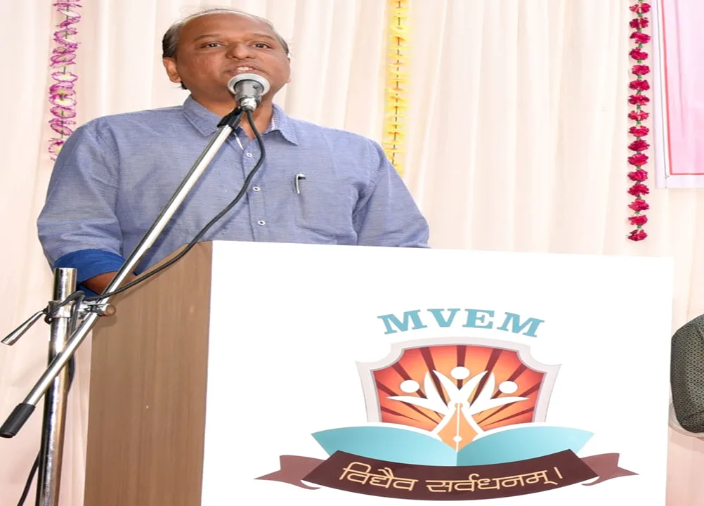 A speaker at a lectern bearing the MVEM crest